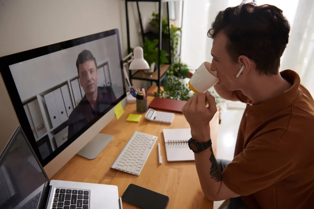 Young man drinking coffee when talking to colleague at online meeting